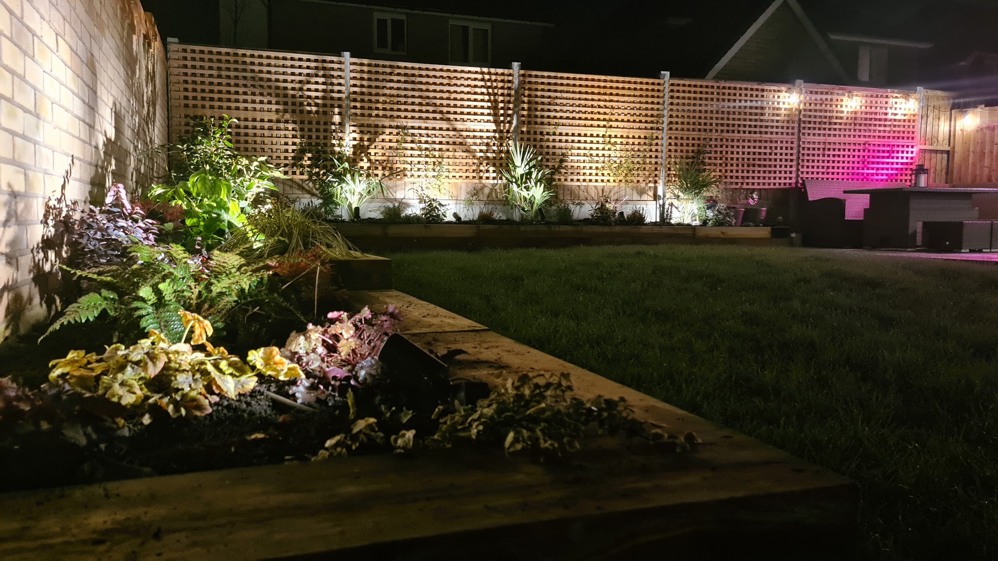 A back garden at night with trellis fence lights and a lit flower bed